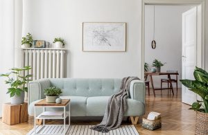 Salotto in stile moderno scandinavo con sofà celeste, tavolino da caffè in metallo bianco e legno, sgabello in legno con sopra una pianta, radiatore bianco in metallo con sopra una mensola e degli oggetti, una mappa incorniciata e appesa alla parete bianca e pavimento a parquet a spina di pesce. Oltre una porta si intravede una sala da pranzo