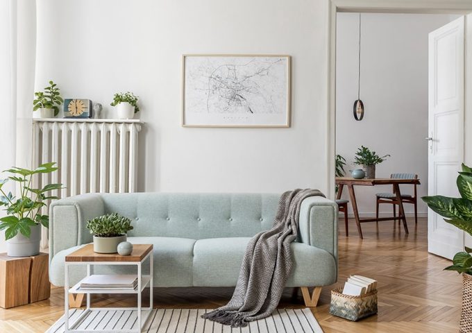 Salotto in stile moderno scandinavo con sofà celeste, tavolino da caffè in metallo bianco e legno, sgabello in legno con sopra una pianta, radiatore bianco in metallo con sopra una mensola e degli oggetti, una mappa incorniciata e appesa alla parete bianca e pavimento a parquet a spina di pesce. Oltre una porta si intravede una sala da pranzo