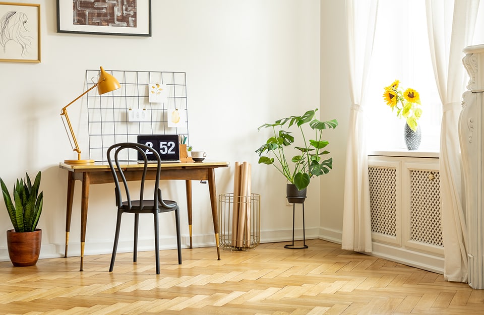 Luminoso angolo domestico con scrivania e sedia in legno, parquet a spina di pesce, piante, una lampada da lettura gialla, dei quadri, e dei soprammobili