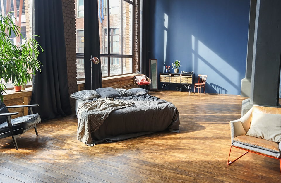 Luminoso e spazioso open space con camera da letto, divani, poltrona, sedia e console. Le pareti sono blu e a mattoncini, il pavimento in parquet rustico, i soffitti sono molto alti e una parete è quasi interamente finestrata