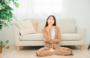 Una donna orientale dall'aria pensierosa è seduta a gambe incrociate sul pavimento in parquet in un salotto con sofà grigio e pianta da interni