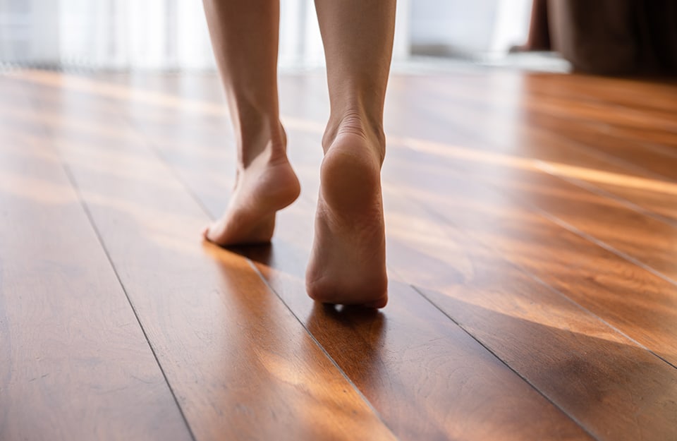 Dettaglio dei piedi scalzi di una donna, visti da dietro, che camminano su un parquet