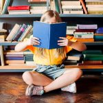 Una bambina bionda seduta a gambe incrociate su un parquet in una stanza con una libreria piena di volumi, sta leggendo un libro che le copre il volto