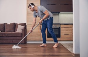 Un uomo scalzo visto di profilo mentre, indossando le cuffie, passa uno spazzolone per pulire il parquet di un open space con cucina e salotto