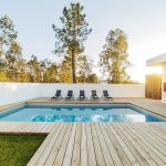 Giardino con decking in legno chiaro e ampia piscina rettangolare. Oltre il muro bianco si vedono degli alti alberi e sulla destra c'è la casa, in stile moderno e minimale