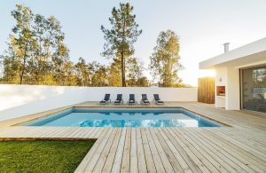 Giardino con decking in legno chiaro e ampia piscina rettangolare. Oltre il muro bianco si vedono degli alti alberi e sulla destra c'è la casa, in stile moderno e minimale