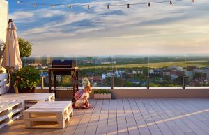 Un bambina piccola con zainetto sulle spalle, accovacciata sul decking di una grande terrazza con arredi realizzati con bancali e affacciata su un panorama verde, con ringhiera trasparente in vetro