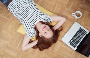 Una ragazza dai capelli rosso-castani e maglietta a righe a maniche corte, fotografata dall'alto, riposa sorridente e a occhi chiusi con le mani incrociate dietro alla nuca su un parquet, accanto a un laptop aperto e una tazza con una bevanda calda