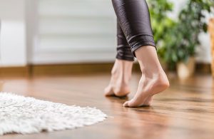 Dei piedi scalzi di donna camminano su un parquet accanto a un tappeto, con una pianta sfocata sullo sfondo
