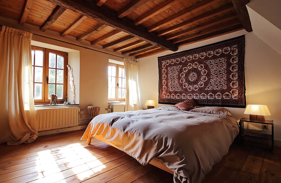 Camera da letto in abitazione rustica con travi a vista. Nella stanza ci sono due finestre, parquet e grande tappeto appeso sopra la testiera del letto