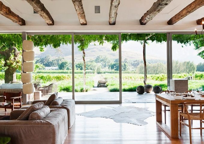 Luminoso salotto in casa di campagna con travi a vista, parete completamente finestrata, diversi divani, tavolo da pranzo in legno e vista sulla campagna