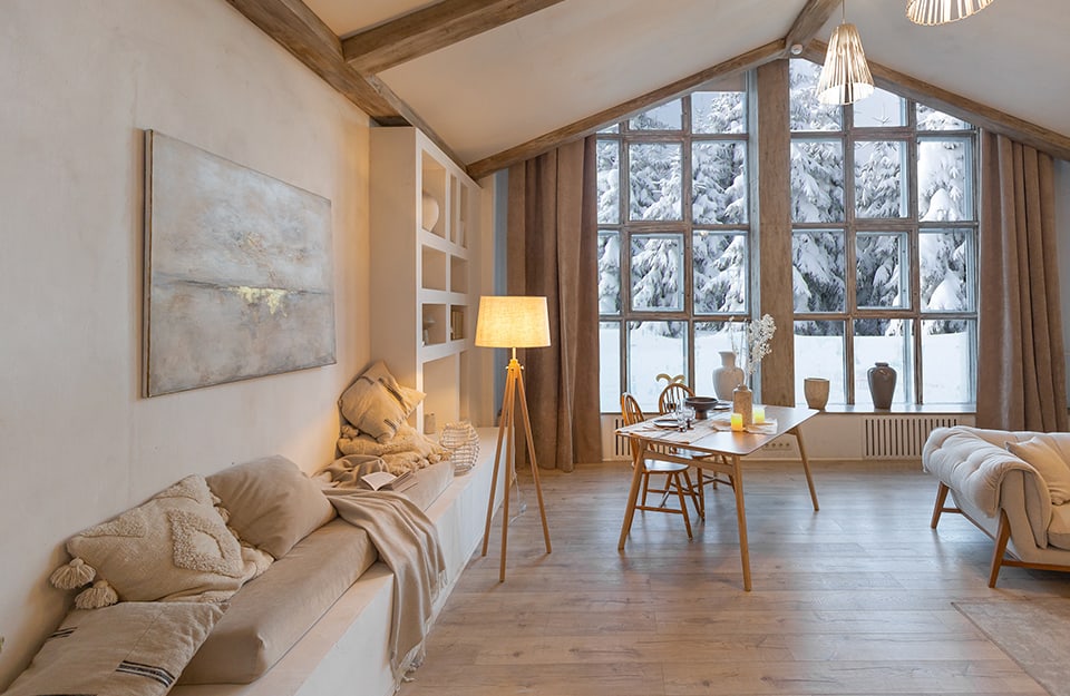 Salotto open-space in stile scandinavo e minimale in una casa di campagna in zona montana, con assi di legno a vista sul soffitto, pavimento in parquet chiaro, divani, scrivania e grande finestra che dà su un panorama boscoso innevato