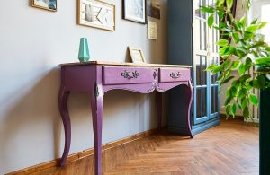 Primo piano di una vecchia consolle dipinta di viola, appoggiata a una parete piena di quadretti, accanto a una vetrinetta vintage dipinta di blu e pavimento in parquet