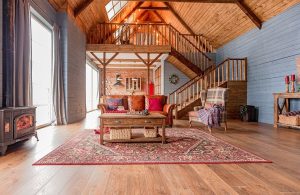 Grande open space di un casa di campagna, probabilmente ricavato da un granaio, tutto in legno (lasciato al naturale per il soffitto, azzurro sulle pareti, e parquet al pavimento) con scale e grande soppalco in legno, stufa antica, sofà e tavolo da caffè sopra un grande tappeto orientale e, sullo sfondo, cucina con una parete di mattoni