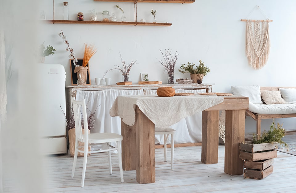 Luminoso open space con cucina, sala da pranzo e salotto in una casa di campagna. La stanza è tutta sui toni del bianco, al centro c'è un massiccio tavolo in legno e si vedono anche un frigorifero bianco, un sofà essenziale in legno, diverse mensole, un parquet bianco come le pareti