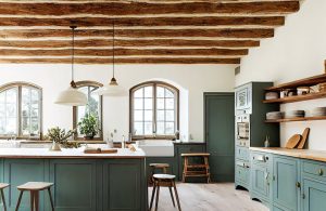 Grande cucina rustica in una spaziosa e luminosa stanza con travi a vista di una casa di campagna. I mobili sono di un verde salvia scuro e c'è una grande isola al centro della stanza