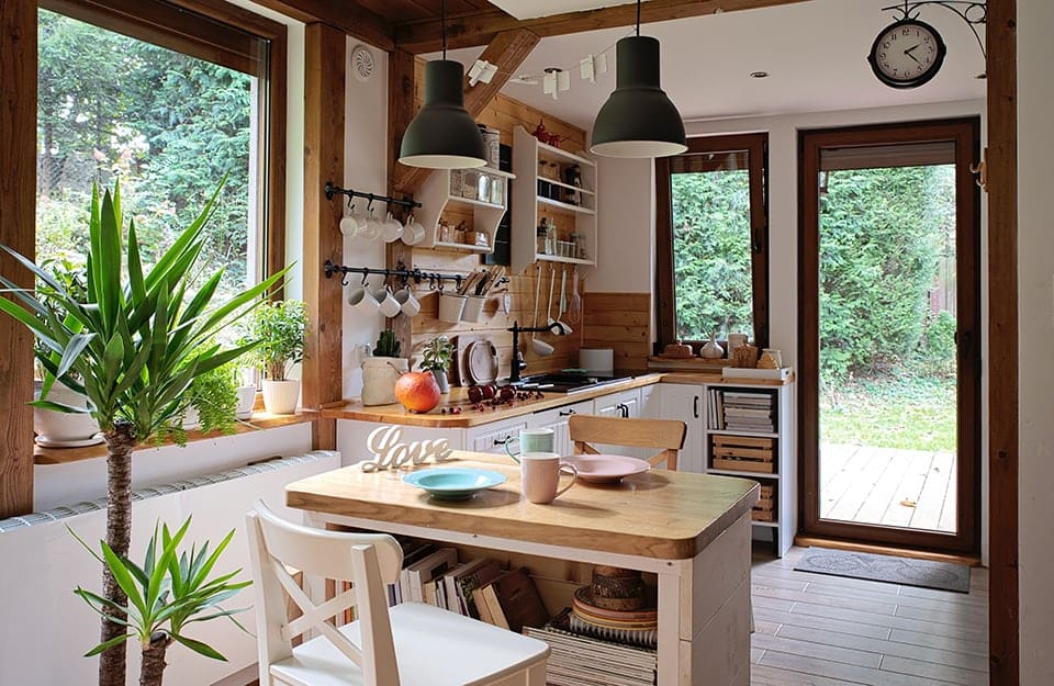 Cucina in stile rustico moderno di una casa in campagna, con angolo cottura bianco e in legno naturale, isola-tavolo, molte finestre, utensili da cucina appesi a vista e diverse piante