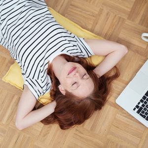 Una ragazza dai capelli rosso-castani e maglietta a righe a maniche corte, fotografata dall'alto, riposa con le mani incrociate dietro alla nuca su un parquet, accanto a un laptop aperto e