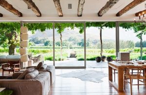 Luminoso salotto in casa di campagna con travi a vista, parete completamente finestrata, diversi divani, tavolo da pranzo in legno e vista sulla campagna