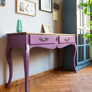 Primo piano di una vecchia consolle dipinta di viola, appoggiata a una parete piena di quadretti, accanto a una vetrinetta vintage dipinta di blu e pavimento in parquet