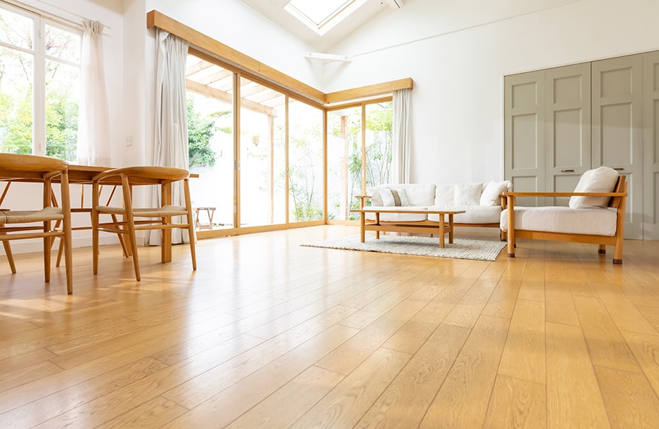 Prospettiva ad altezza pavimento di un soggiorno luminoso ed essenziale, con pochi mobili (tavolo con sedie, divano, poltrona, tappeto e tavolino da caffè), con parquet lucido color miele e grande finestrone d'angolo