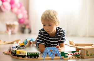 Un bambino biondo sdraiato sul parquet sta giocando con dei trenini su un binario di legno