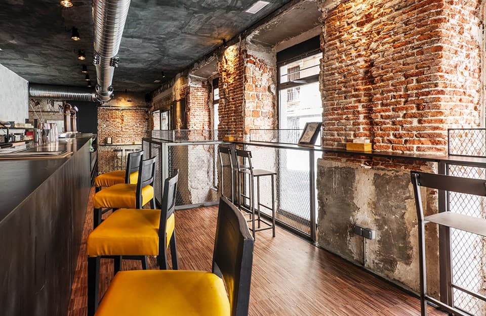 Un bar in stile industriale con grande bancone, tubi a vista, muro in mattoncini, parquet industriale e sedute con imbottitura gialla
