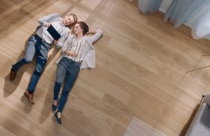 Un ragazzo e una ragazza fotografati dall'altro sdraiati confortevolmente sul parquet del loro salotto mentre guardano insieme lo schermo di un tablet