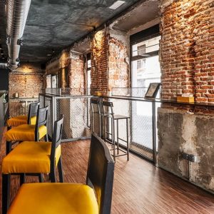 Un bar in stile industriale con grande bancone, tubi a vista, muro in mattoncini, parquet industriale e sedute con imbottitura gialla