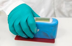 La mano guantata di un tecnico in tuta bianca sta usando un glossmetro su un campione di legno verniciato