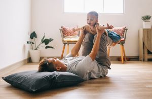 Un padre sdraiato su un pavimento in parquet, con la testa appoggiata a un cuscino sta giocando con su figlio piccolo sollevandolo in aria come fosse un aeroplano