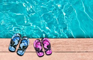 Vista dall'alto di due paia di infradito di colori diversi (azzurre e magenta) lasciate sulle assi in legno del decking a bordo piscina