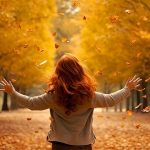 Una donna coi capelli rossi, vista di spalle, apre le braccia con gioia al paesaggio autunnale che ha davanti, pieno di alberi dalle foglie ingiallite e rossastre che ricoprono anche la strada