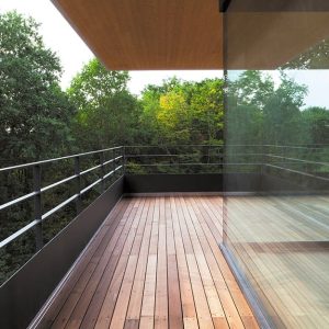 Terrazza esterna con pavimento in decking, ringhiere in metallo, e grande parete vetrata che separa l'interno dall'esterno, immerso nel verde degli alberi