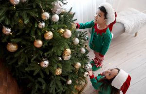 Vista dall'alto, di tre quarti, di un bambino e una bambina in pigiama e con in testa il cappello di Babbo Natale che stanno addobbando un albero di Natale con palline dorate e argentate