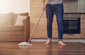 Un uomo scalzo, del quale si vede solo dal busto in giù, sta passando lo straccio sul parquet in una casa con mobili moderni in legno