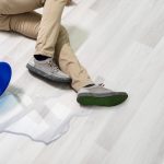 Un uomo caduto a terra (di lui si vedono solo le gambe) su un pavimento in parquet scivoloso per via di una piccola pozza d'acqua rovesciatasi da un secchio