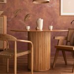 Stanza con parete marrone-rossiccia con effetto limewash, tavolo in legno circolare, sedie in legno in stile scandinavo, armadietto con ante in vimini, pavimento in parquet