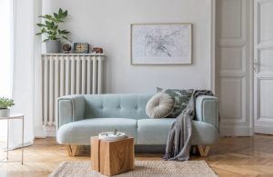 Salotto moderno con sofà celeste dalle linee tondeggianti, pareti bianche, con quadro e termosifone bianco in ghisa, blocco di legno come tavolino da caffè e parquet a spina di pesce