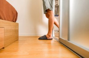 Le gambe di un uomo in ciabatte che esce dalla porta scorrevole di un bagno e, camminando sul parquet, si avvicina al letto