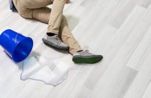 Un uomo caduto a terra (di lui si vedono solo le gambe) su un pavimento in parquet scivoloso per via di una piccola pozza d'acqua rovesciatasi da un secchio
