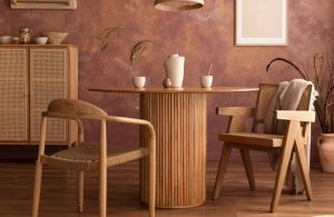 Stanza con parete marrone-rossiccia con effetto limewash, tavolo in legno circolare, sedie in legno in stile scandinavo, armadietto con ante in vimini, pavimento in parquet