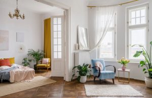 Luminoso open space con pareti e soffitto bianchi, parquet scuro, letto, poltrona, tappeto, porta a vetri e molte piante, con mobili dai colori d'accento accesi
