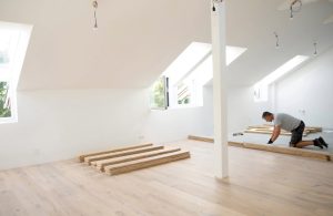 Un posatore di parquet, visto da qualche metro di distanza, sta installando un pavimento in legno in una mansarda di un nuovo edificio. Le mura sono bianche e si vedono dei cavi elettrici scendere dal soffitto spiovente
