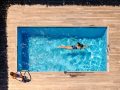 Vista dall'alto di una piscina domestica all'aperto, con una donna che fa il bagno e un decking in legno chiaro che circonda la piscina