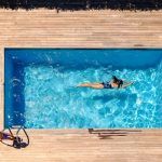 Vista dall'alto di una piscina domestica all'aperto, con una donna che fa il bagno e un decking in legno chiaro che circonda la piscina