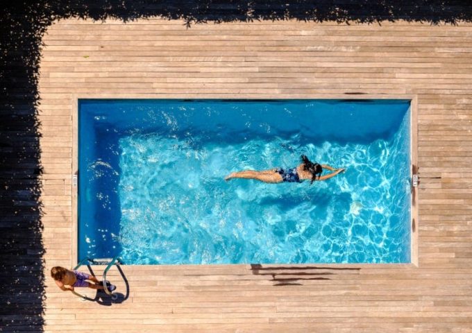 Vista dall'alto di una piscina domestica all'aperto, con una donna che fa il bagno e un decking in legno chiaro che circonda la piscina