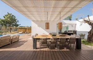 Un'area patio esterna con un decking in legno. Al centro, c'è un grande tavolo da pranzo con otto sedie disposte intorno. Sopra il tavolo, c'è una pergola con un tetto a doghe che fornisce un'ombra parziale. Due lampade a sospensione in vimini pendono dalla pergola sopra il tavolo. A sinistra, ci sono due panchine in legno che si fronteggiano, con vista su una piscina e una pergola in legno sullo sfondo. L'area è circondata da vegetazione, tra cui un grande albero sul lato destro dell'immagine. Il patio è adiacente a un edificio moderno bianco con grandi finestre