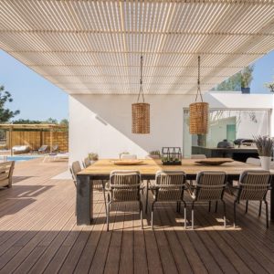 Un'area patio esterna con un decking in legno. Al centro, c'è un grande tavolo da pranzo con otto sedie disposte intorno. Sopra il tavolo, c'è una pergola con un tetto a doghe che fornisce un'ombra parziale. Due lampade a sospensione in vimini pendono dalla pergola sopra il tavolo. A sinistra, ci sono due panchine in legno che si fronteggiano, con vista su una piscina e una pergola in legno sullo sfondo. L'area è circondata da vegetazione, tra cui un grande albero sul lato destro dell'immagine. Il patio è adiacente a un edificio moderno bianco con grandi finestre