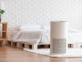 Un purificatore d'aria bianco in primo piano in una camera da letto con pavimento in parquet, pareti bianche con mattoni a vista e letto con struttura in pallet